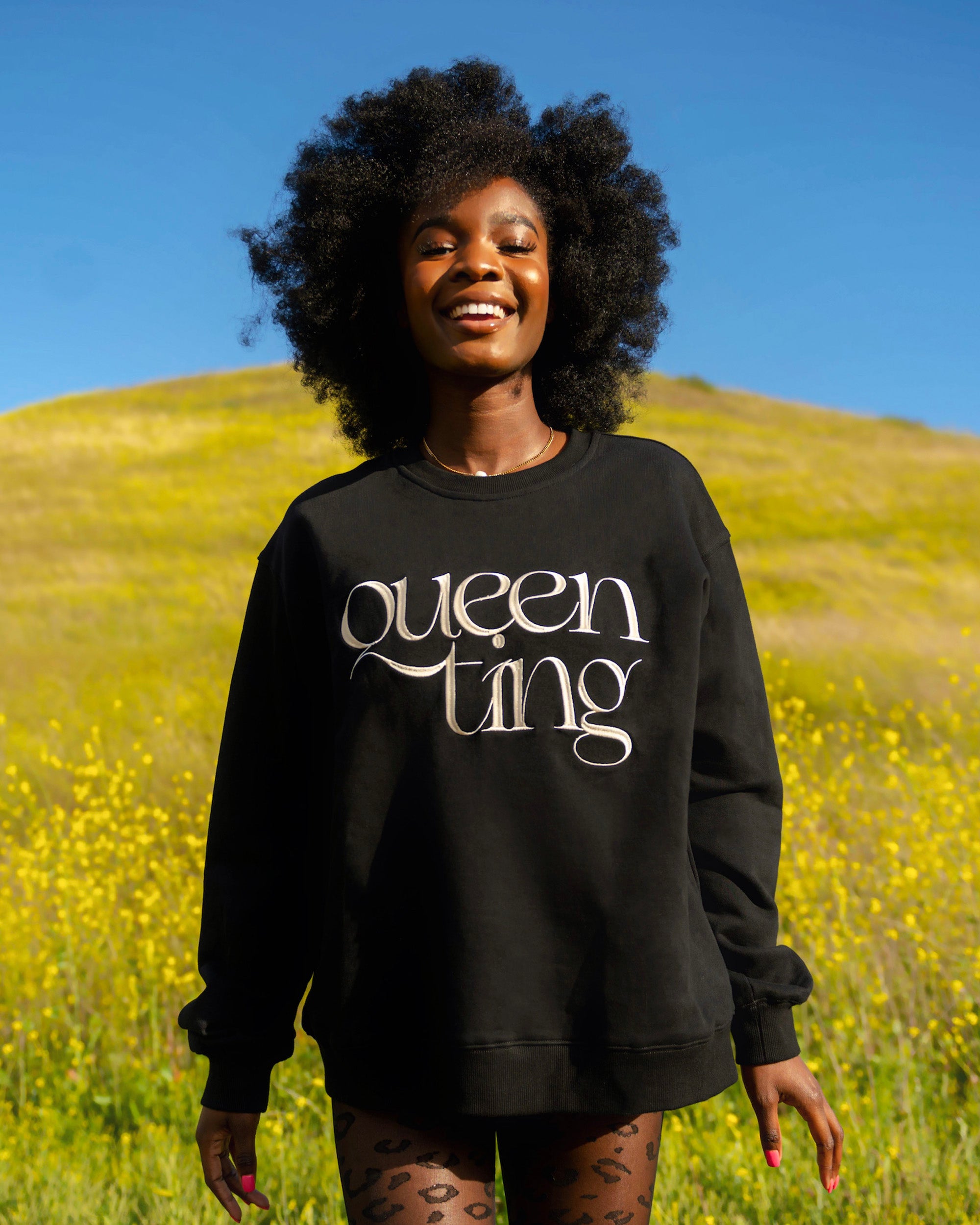 Beautiful black female model with afro hair smiling on a grass hill wearing the Queen Ting embroidery sweatshirt by SHOLAYIDÉ