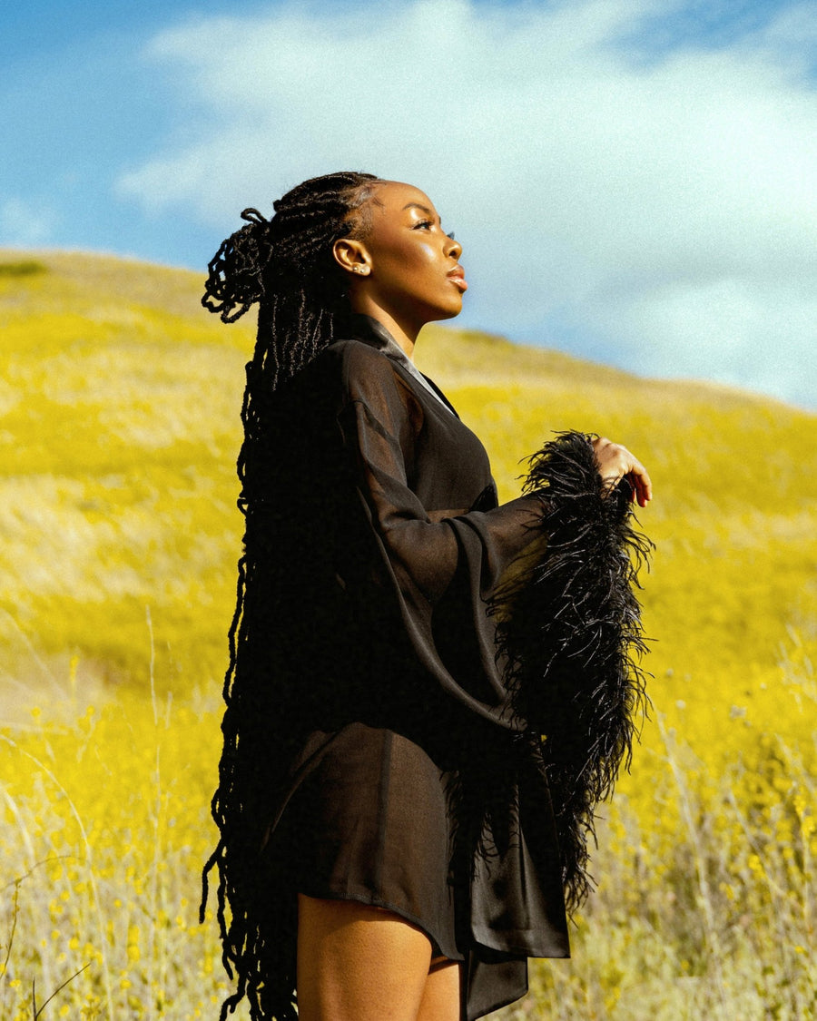 Black female model posing on a grassy hill wearing the EDEN feather trim robe by SHOLAYIDÉ