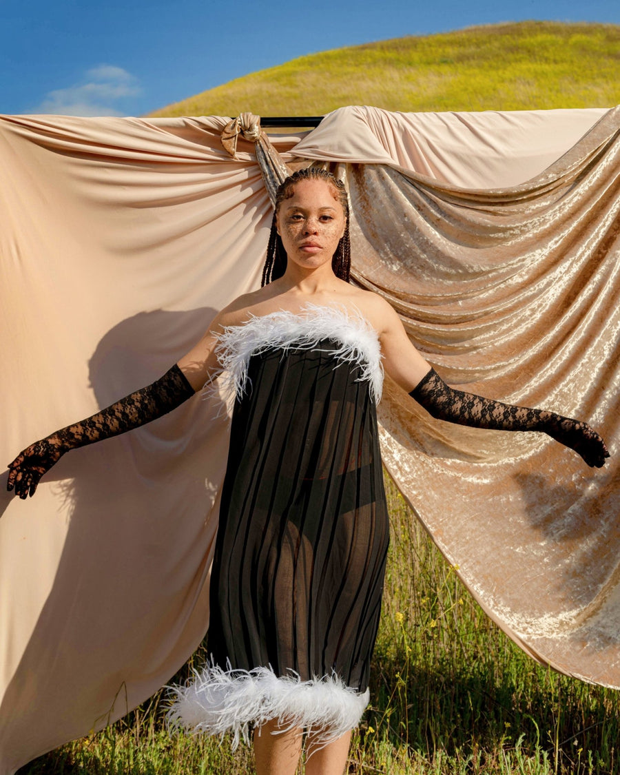 Black female model with freckles posing on a grassy hill wearing EVA feather trim dress by SHOLAYIDÉ.