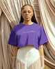 Black female model with freckles posing in front of draped curtains wearing FAVORED purple cropped tee by SHOLAYIDÉ.