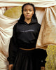 Black female model posing in front of draped curtains wearing HEAVENLY cropped hoodie with embroidery by SHOLAYIDÉ.